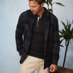 Man wearing a black and gray Reggie Flannel Shirt over a black henley, styled casually with beige pants indoors.