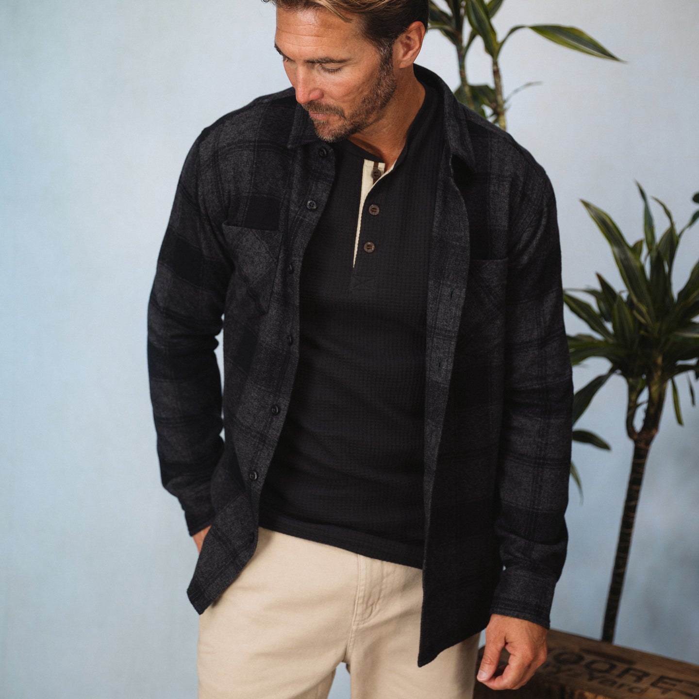 Man wearing a black and gray Reggie Flannel Shirt over a black henley, styled casually with beige pants indoors.