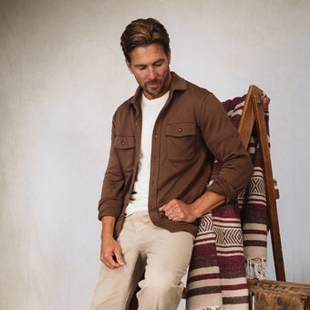 Man in a brown overshirt and beige pants seated beside a patterned blanket draped over a wooden ladder chair