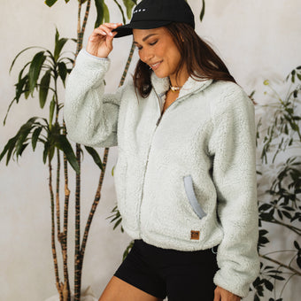 Woman wearing a cozy fleece jacket and black cap, smiling indoors among tropical plants