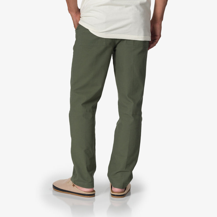 Back view of a man wearing Matthew Twill Pant in thyme green, paired with beige surf-inspired sandals facing right.
