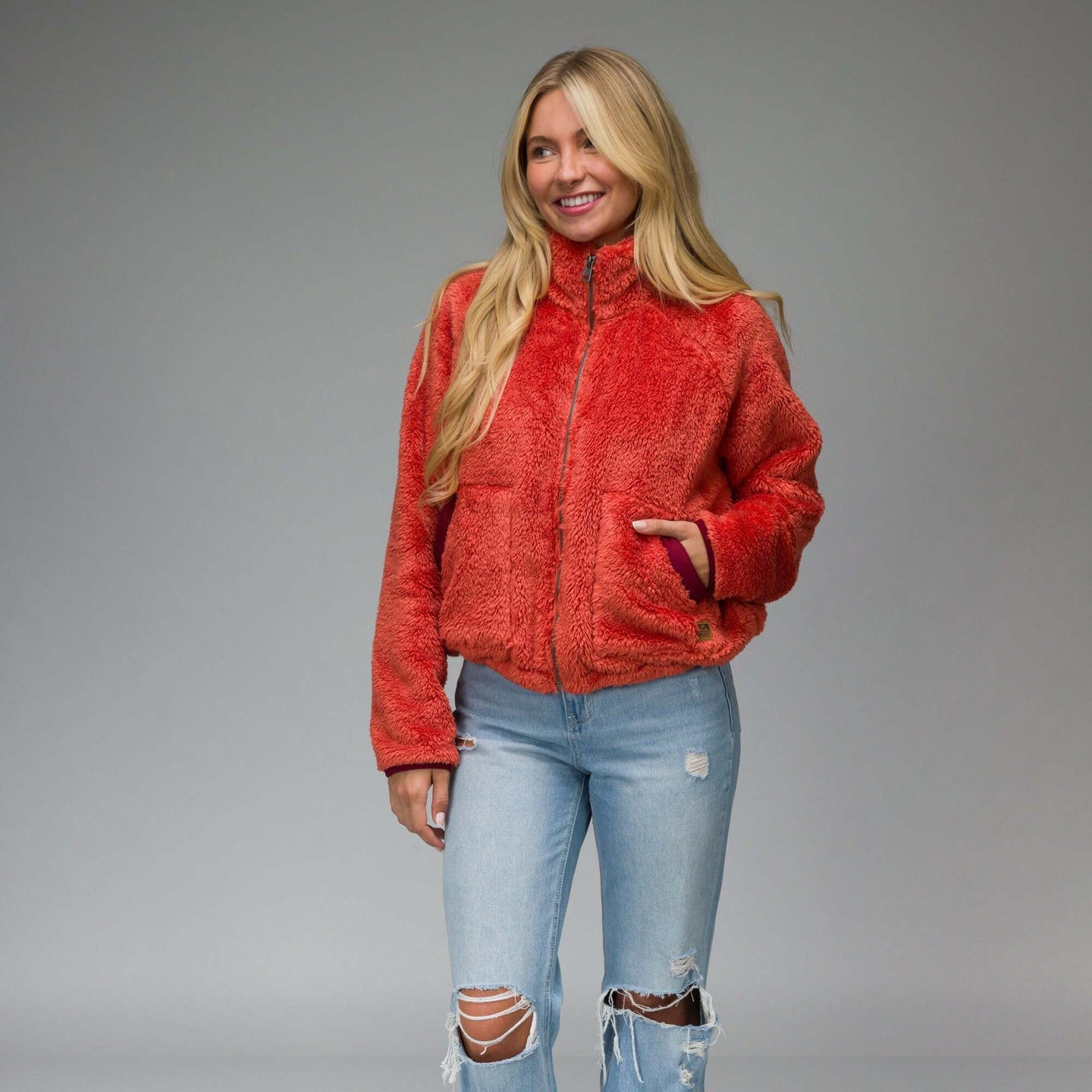 Woman wearing a bright chili red Pilot Sherpa Jacket facing front with hands in pockets, showing its cozy textured fabric.