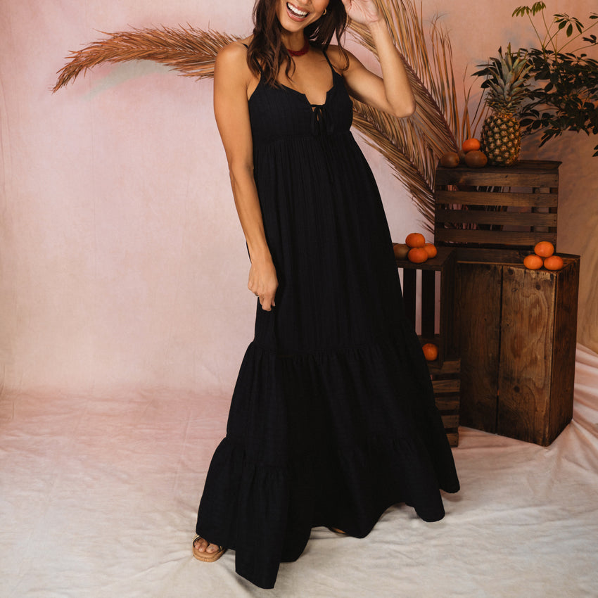 Woman wearing a black Yuni Maxi Dress with a deep notched neckline and tie detail, styled in a beach-inspired setting.