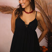 Woman wearing a black Yuni Maxi Dress with a deep notched neckline and delicate tie detail, smiling indoors.