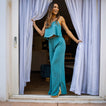 Woman wearing teal Ollie Pant standing in doorway with white curtains, showcasing breathable beach-inspired comfort apparel