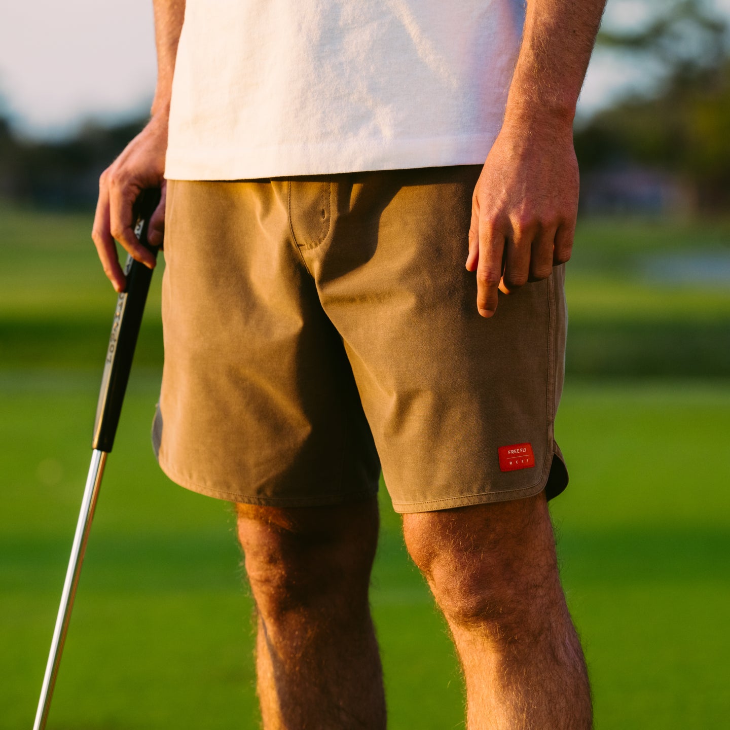 Men wearing Men's Reverb Short Unlined – 7" - Coconut standing on grass holding golf club, casual activewear style