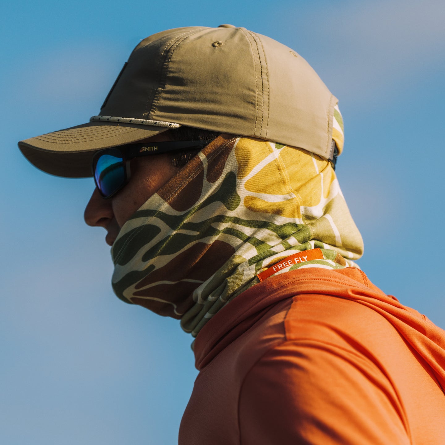 Man facing left wearing Bamboo Lightweight Sun Mask - Floral Camo with tan hat, sunglasses, and orange shirt in bright sunlight