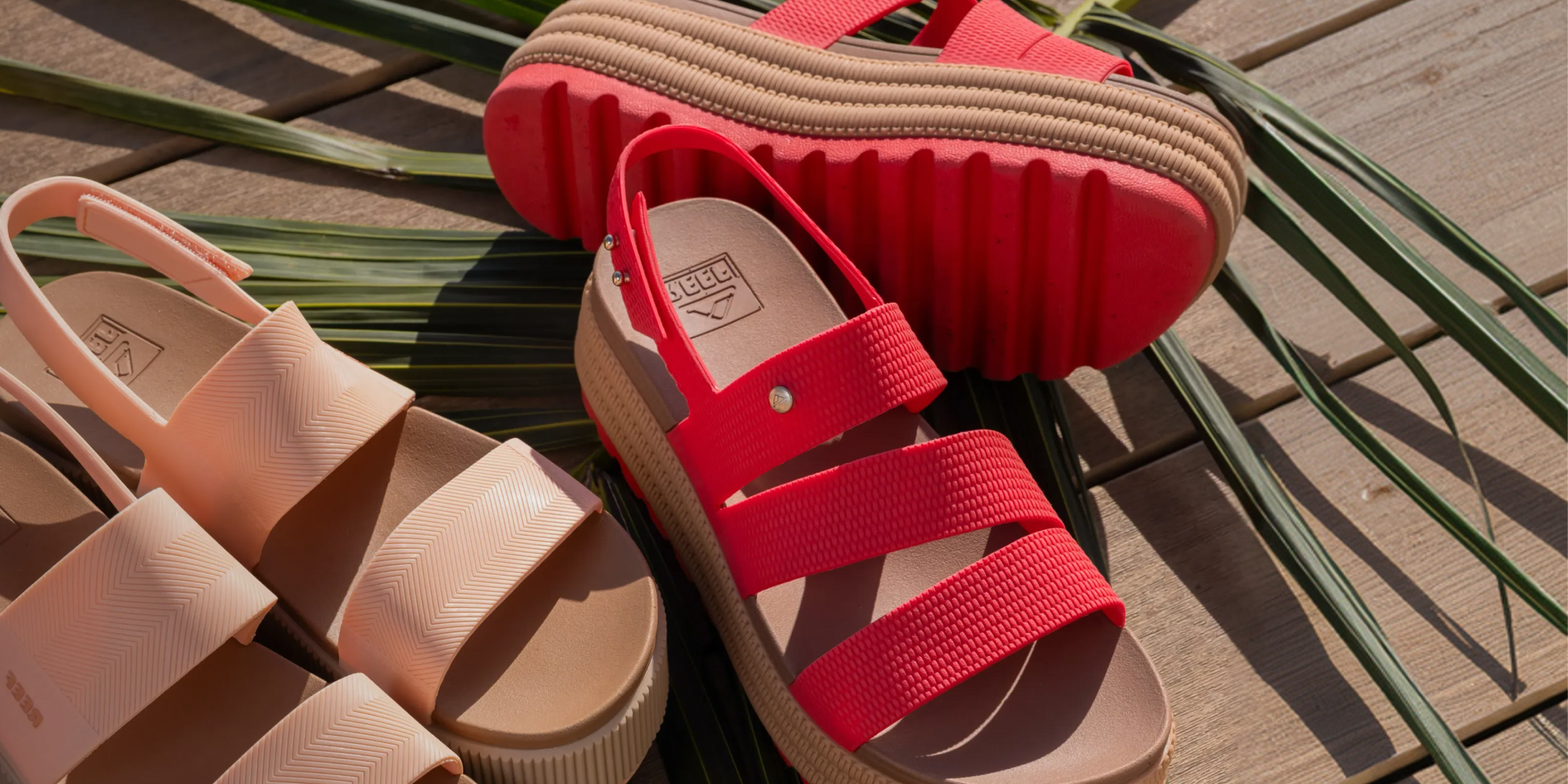 Close-up of right-facing red platform sandals with textured straps and cushioned footbeds resting on wooden surface with palm leaves nearby