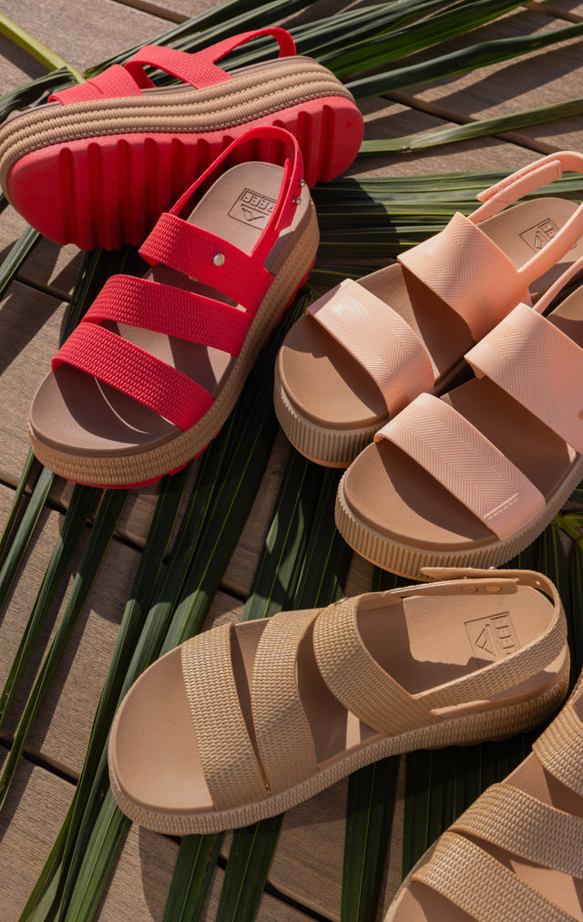 Top view of left-facing platform sandals in red, beige, and blush with cushioned footbeds and textured straps on palm leaves