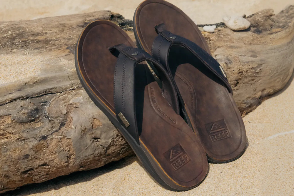 Pair of brown REEF sandals angled upward on sandy beach with textured footbeds and durable straps near driftwood log