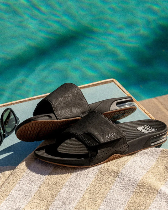 Side view of black REEF slides facing right with textured soles and cushioned footbeds by poolside on towel surface