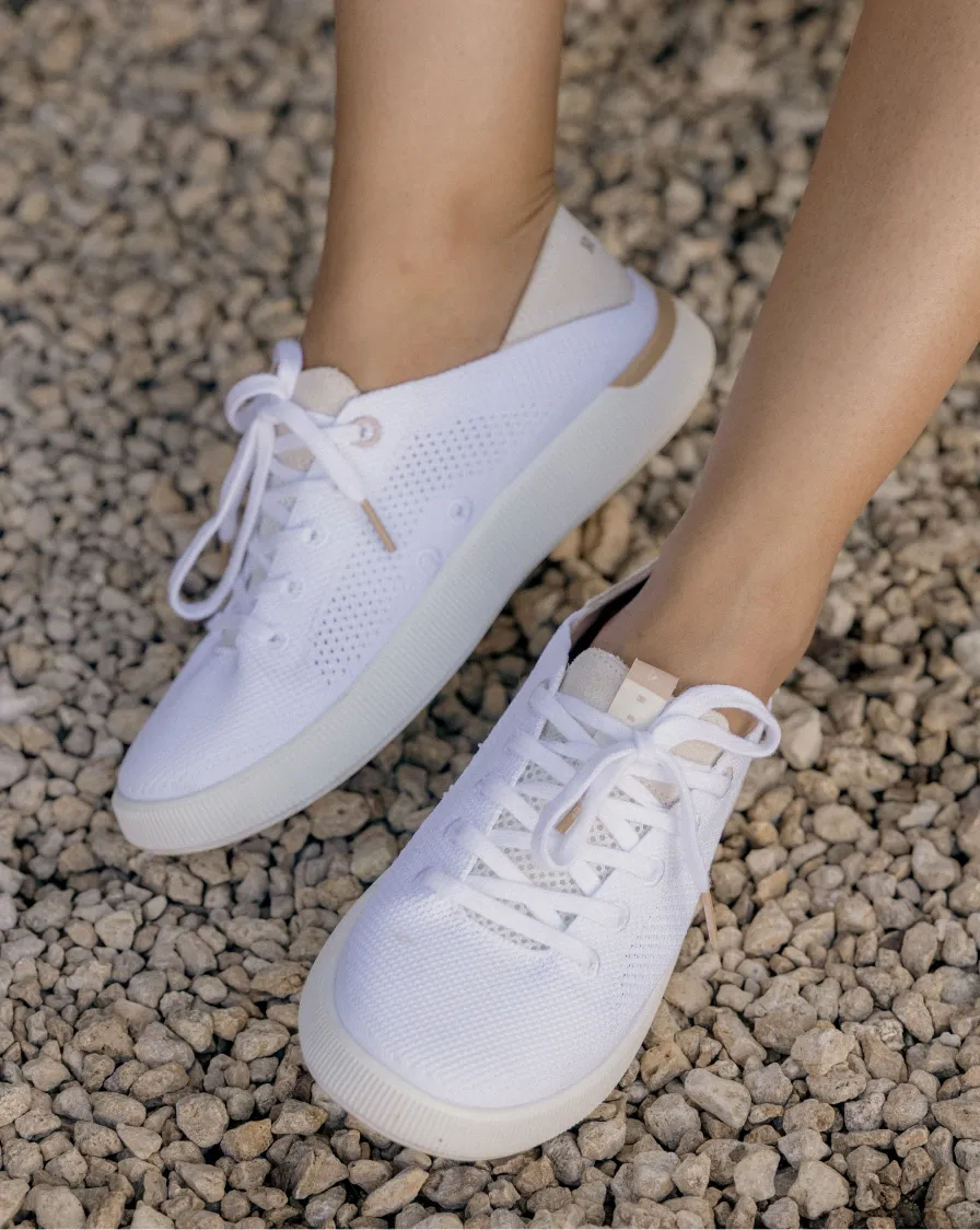 Top-down view of feet wearing right-facing white sneakers with mesh uppers and cushioned soles on gravel ground
