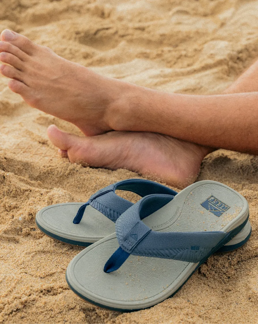 Blue REEF flip flops with textured footbeds resting on sand near relaxed bare feet, showcasing beach-ready comfort