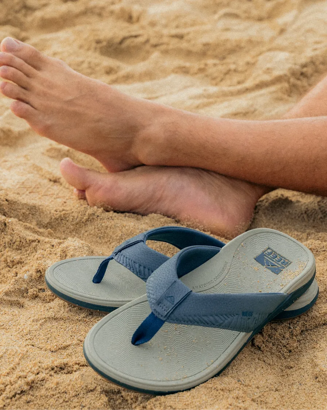 Blue REEF flip flops with textured footbeds resting on sand near relaxed bare feet, showcasing beach-ready comfort