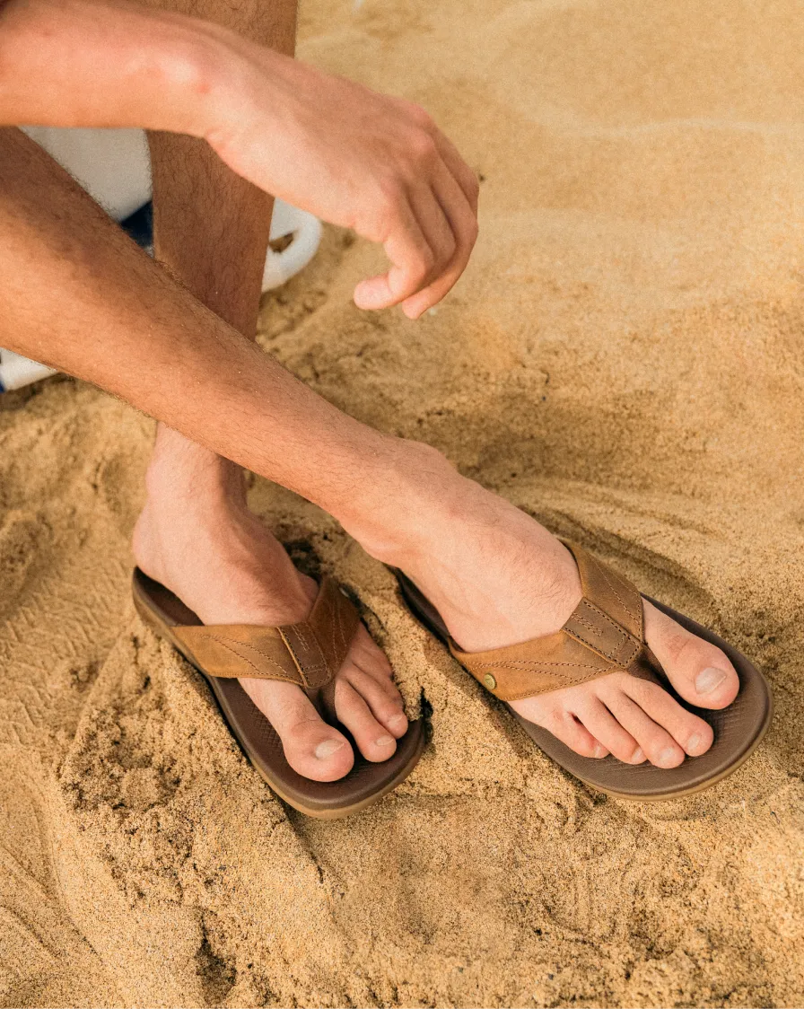 Close-up of feet wearing brown leather flip flops facing forward with cushioned footbeds on sandy beach surface