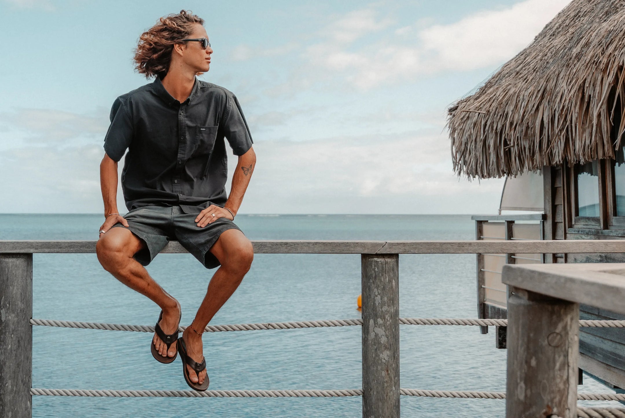 Side view of man wearing black flip flops facing left while sitting on wooden railing by ocean and beach hut