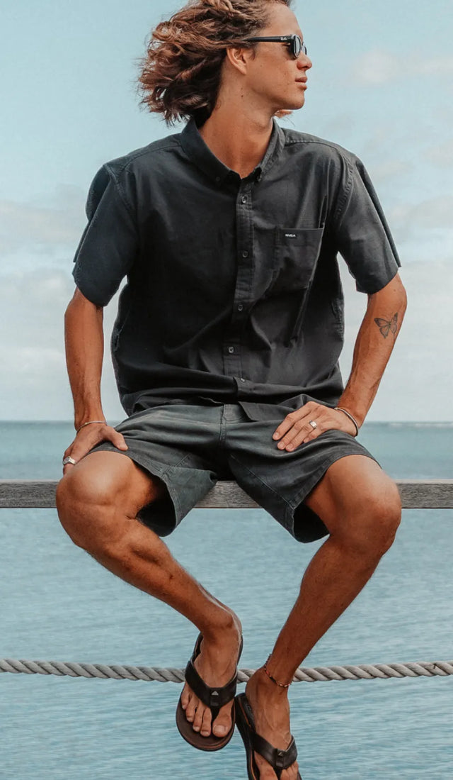 Man seated on railing by the ocean wearing black shirt, dark shorts, and black flip flops facing forward with relaxed posture