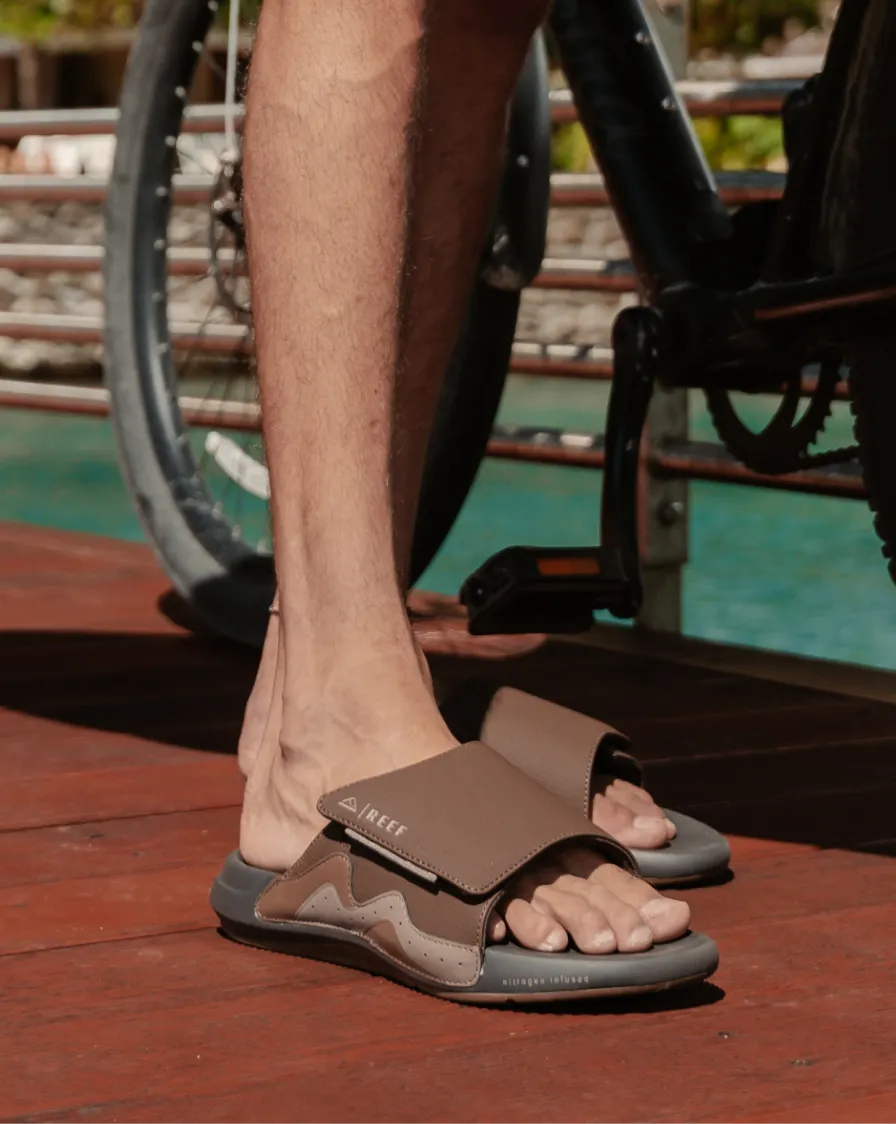 Close-up of left foot wearing taupe slide sandal with adjustable strap and cushioned sole standing on wooden deck near bicycle wheel