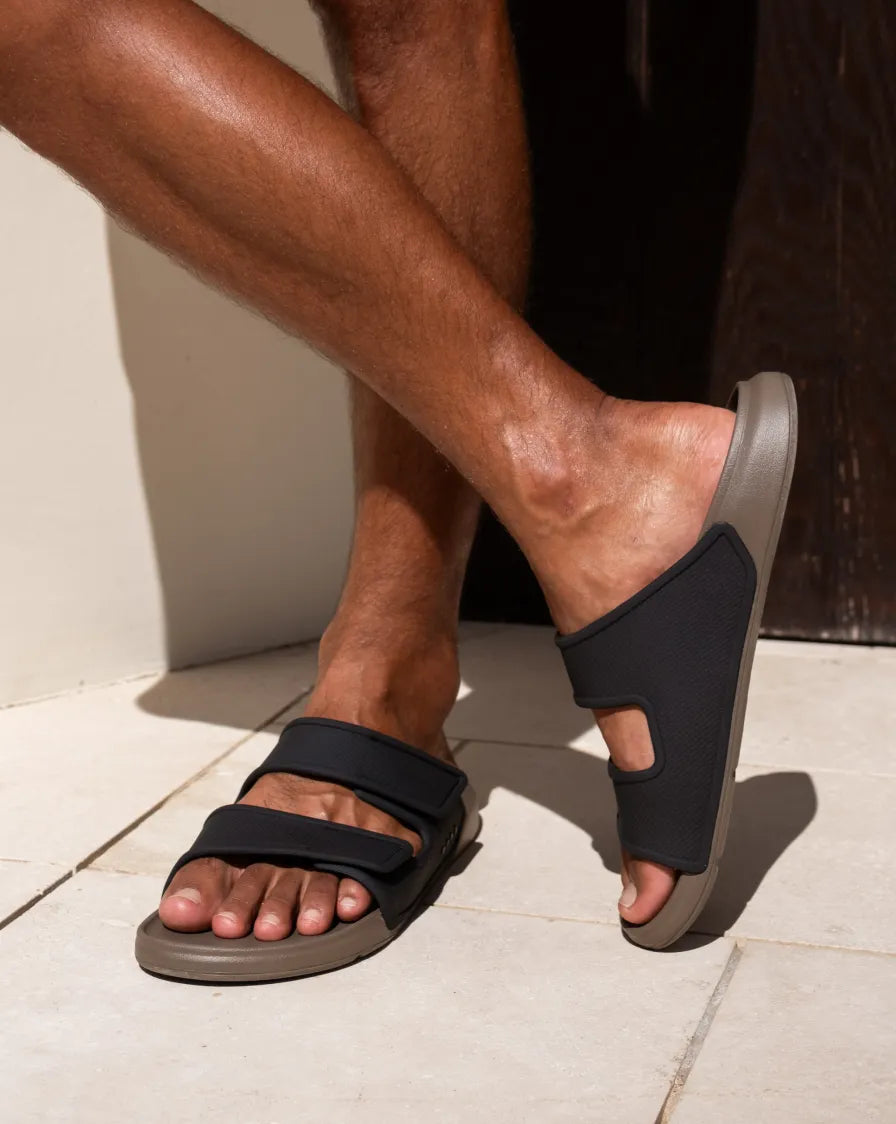 Right-facing black and gray REEF slides on a bare foot with adjustable straps and cushioned, water-friendly footbeds outdoors