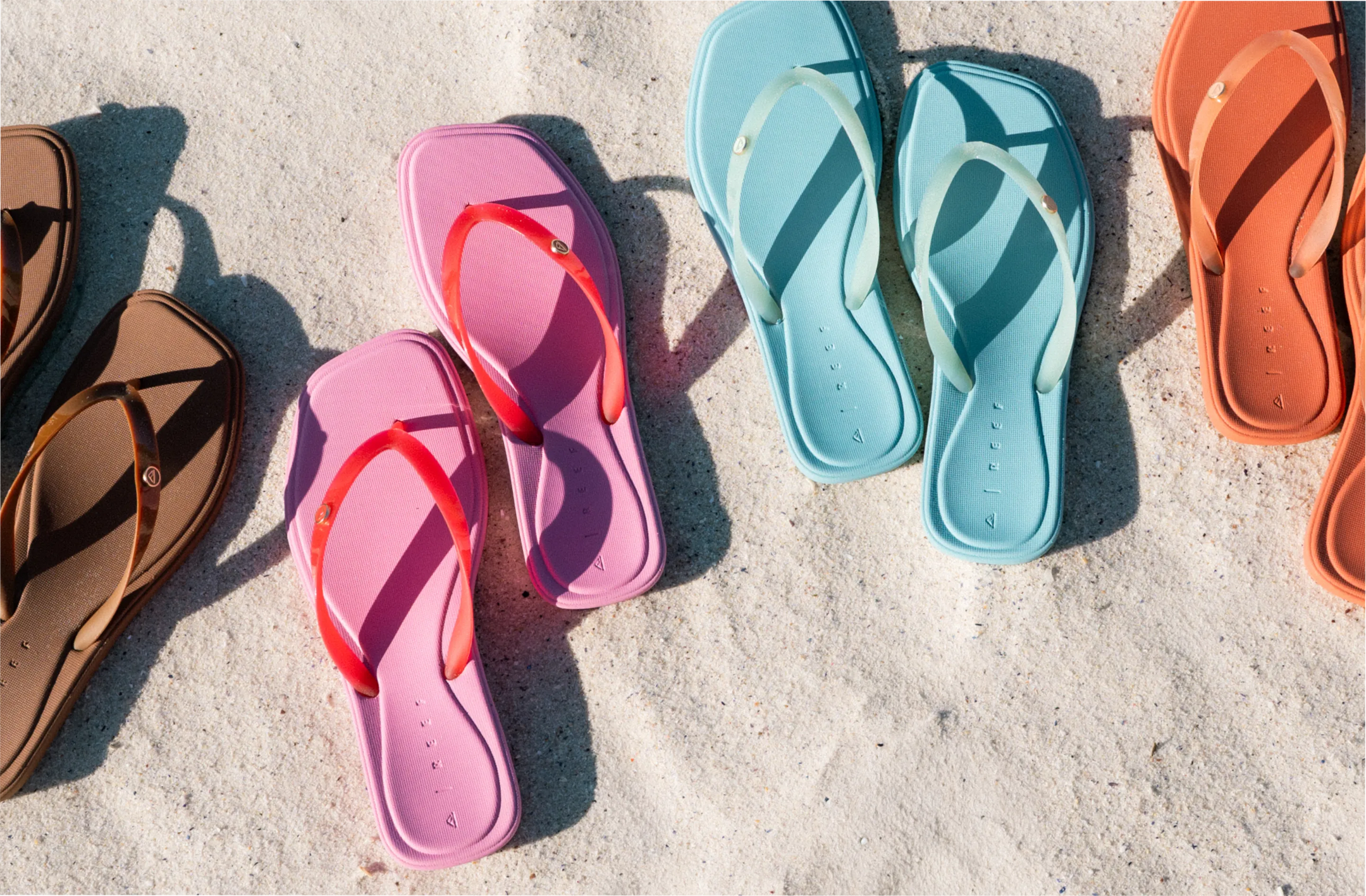 Four pairs of colorful flat sandals in brown, pink with red straps, light blue, and orange, arranged on white sand.