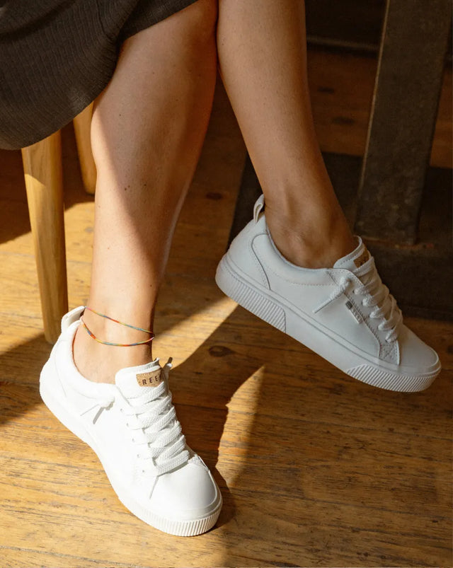 Pair of right-facing white REEF sneakers with cushioned soles and laces, shown from side on wood floor with model's legs visible