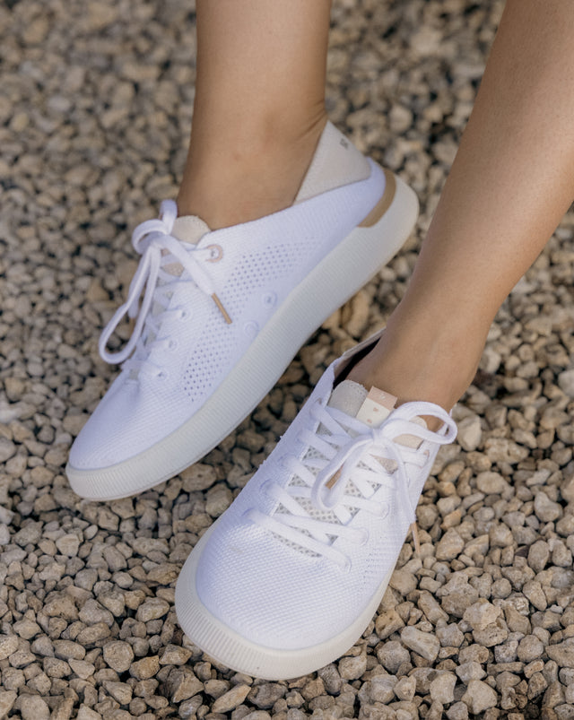 Close-up of feet wearing white REEF sneakers facing forward with breathable knit uppers on rocky ground background