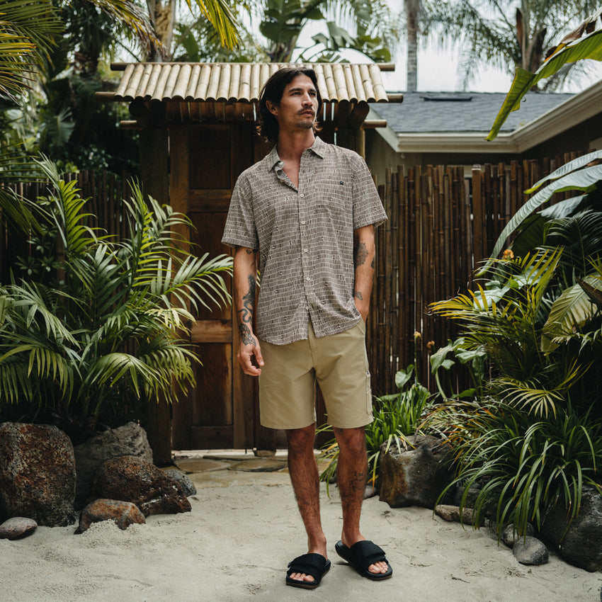 Man wearing Ferris Classic Shirt in neutral tones standing outdoors by tropical plants, styled for a relaxed beach lifestyle.