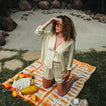 Woman wearing Ellie Button Up cream striped shirt kneeling on beach towel with fruits nearby, shaded by her hand