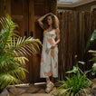 Woman wearing Belladona Dress with floral print, standing outdoors near wooden door and bamboo fence, sandals visible facing forward.