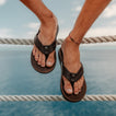 Leather Offshore men's sandals in dark brown, front facing close-up showing padded straps and cushioned footbed by the ocean.