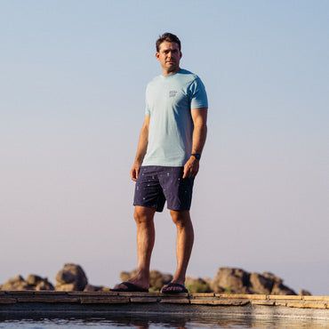 Man wearing La Loca Tee in light blue, standing outdoors in beach-inspired casual apparel with sandals facing forward.
