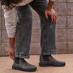 Man wearing gray jeans puts on right-facing Evo mid-cut black leather footwear with cushioned sole in outdoor setting