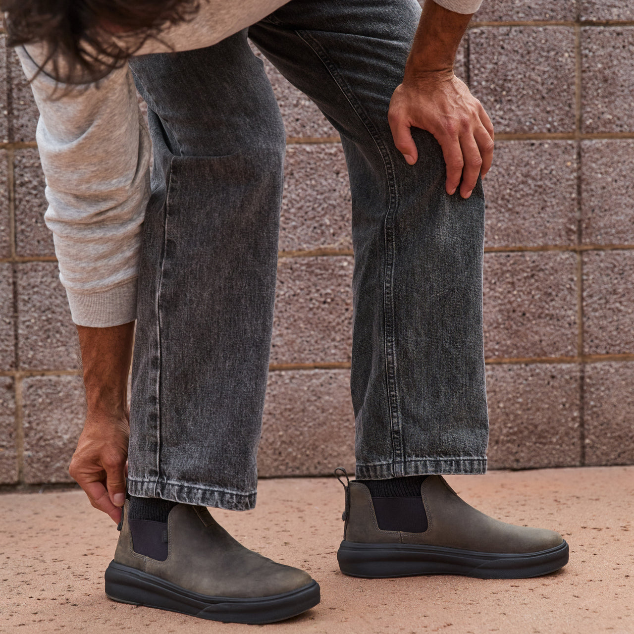 Man wearing gray jeans puts on right-facing Evo mid-cut black leather footwear with cushioned sole in outdoor setting