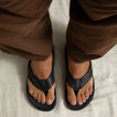 Top view of adult men's Phantom Lux black flip flops with anatomical arch support and soft cushioning, worn with brown pants.