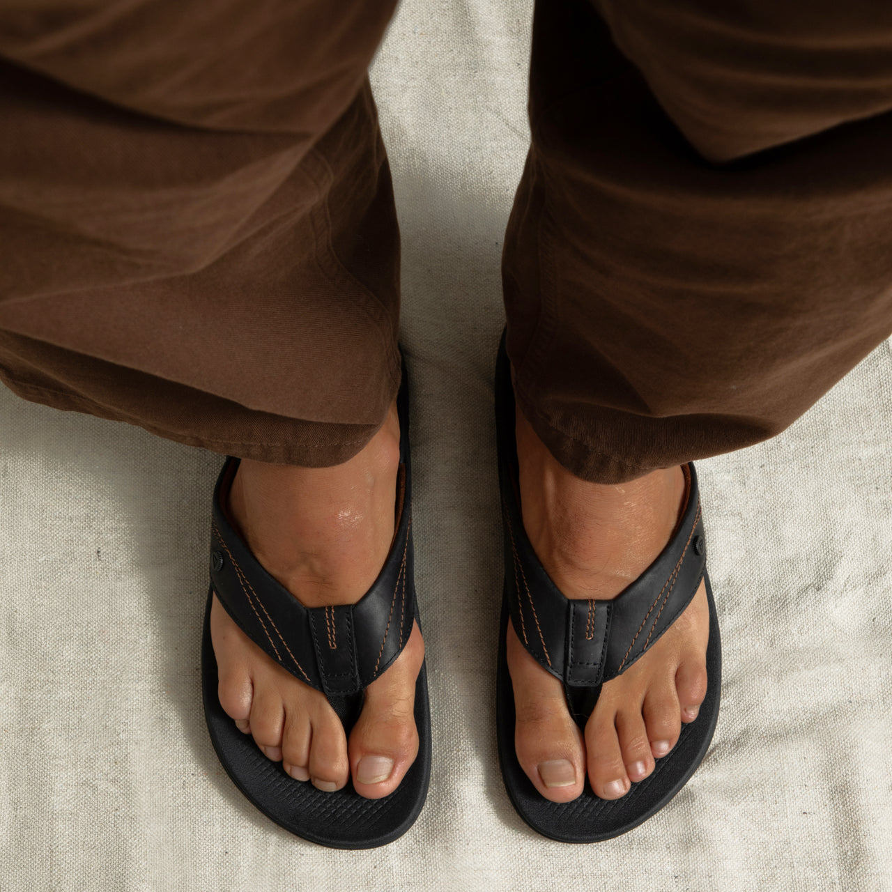 Top view of adult men's Phantom Lux black flip flops with anatomical arch support and soft cushioning, worn with brown pants.