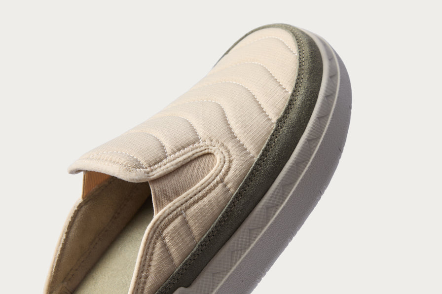 Close-up of beige slip-on sneaker facing left showing quilted fabric upper, cushioned collar, and textured sole edge.