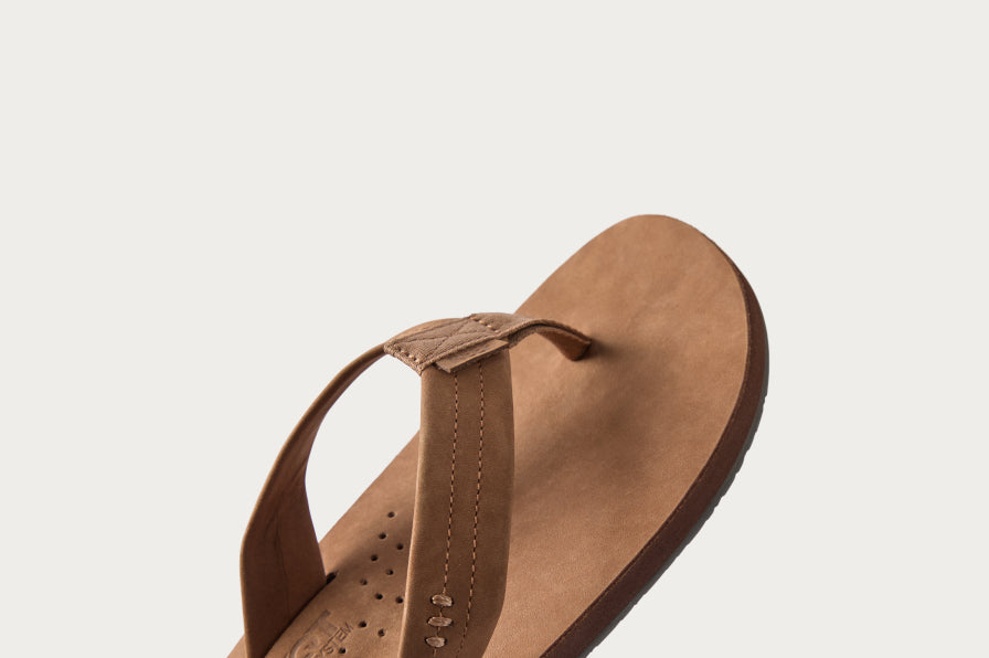 Close-up of right brown leather flip flop facing left, showing cushioned strap and smooth footbed with subtle perforations.