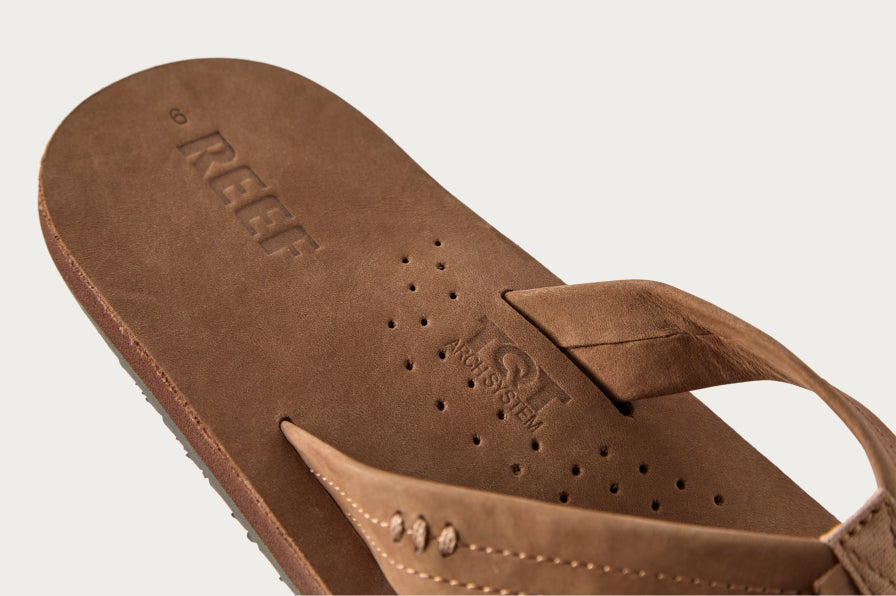Close-up of brown leather flip flop facing left showing perforated footbed, cushioned strap, and embossed REEF logo.