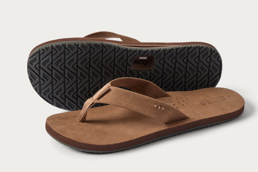Pair of brown leather flip flops, one facing left showing textured sole, the other top-side up with perforated footbed and cushioned strap