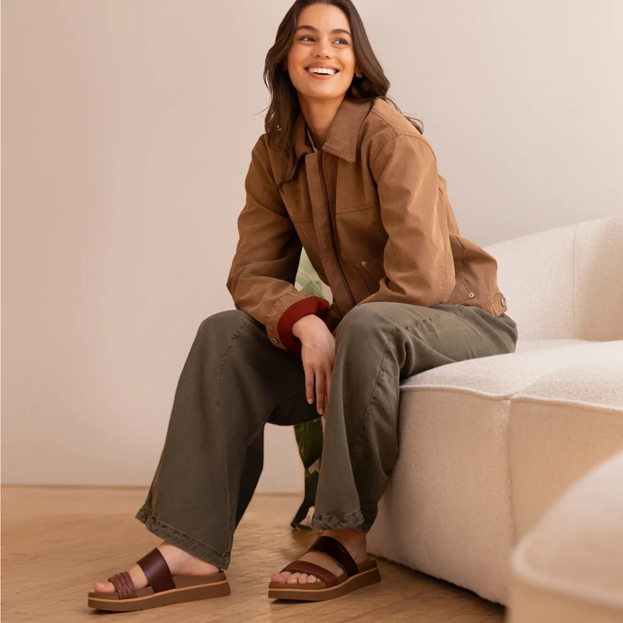 Woman sitting on beige couch wearing brown Vista Hi 2 sandals facing forward, showing sandal straps and platform sole.