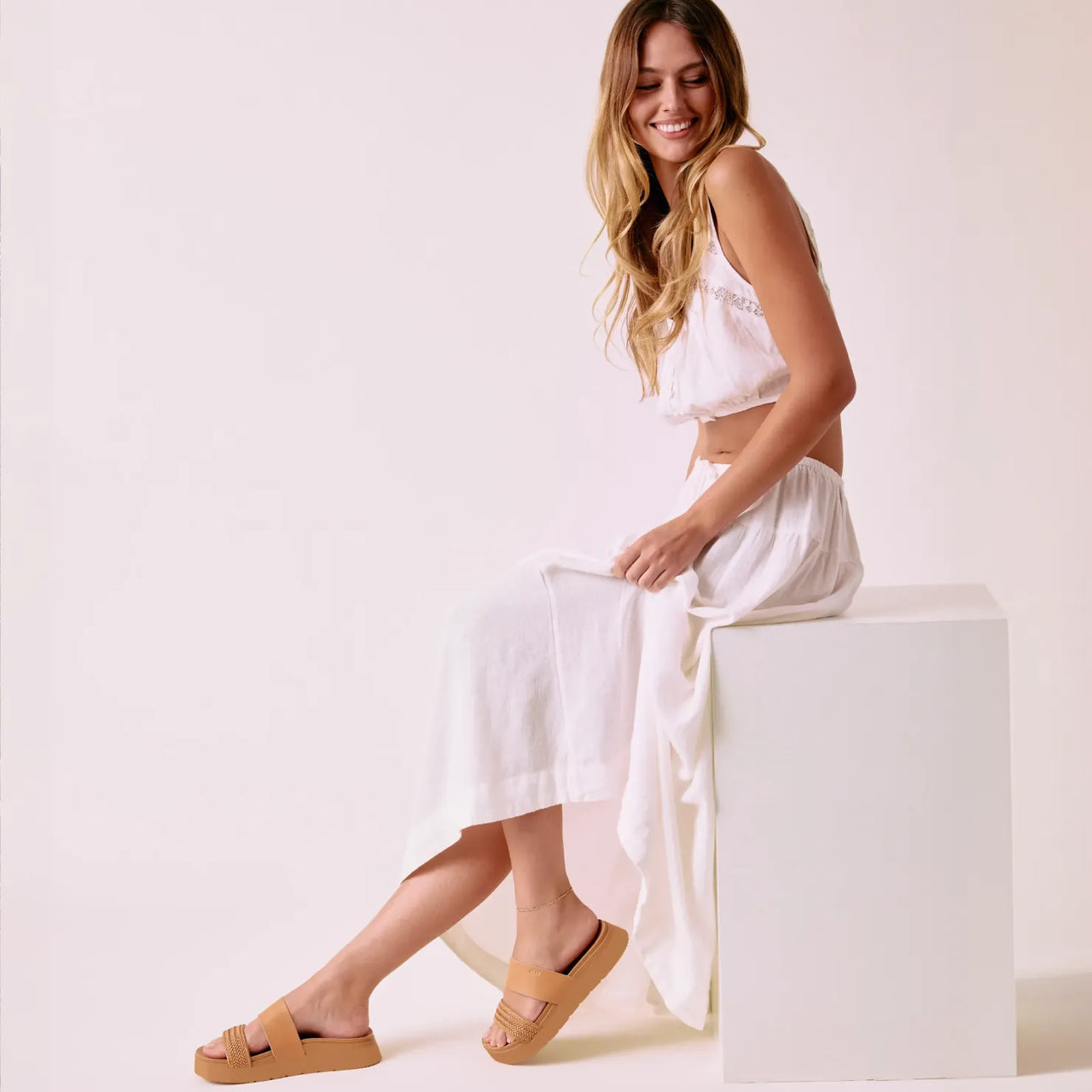 Woman sitting on white block facing left wearing Vista Hi 2 tan sandals with enhanced arch support and cushioned platform