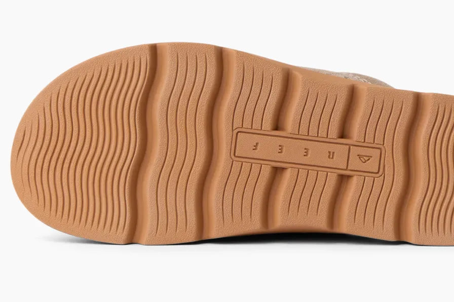 Close-up of the textured tan rubber sole of a sandal facing upward showing wave-like traction and recessed REEF logo plate
