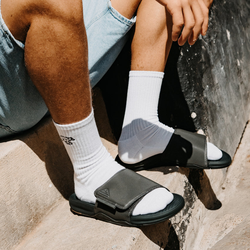Phantom Slab black slides facing right with cushioned footbeds on a rocky beach