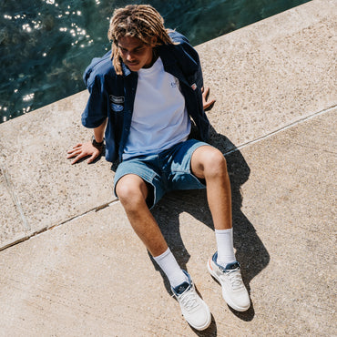 Shoreline Del Mar white sneakers facing toward the lower right as a man sits beside the water