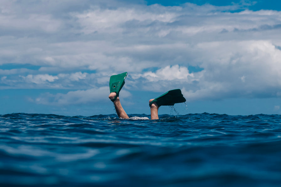 Person’s legs with green swim fins visible above deep blue ocean water under partly cloudy sky