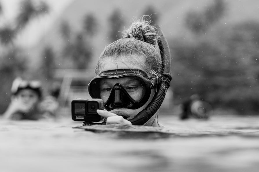 Black and white photo of a person wearing a snorkel mask and holding an action camera while floating in water near blurred figures.