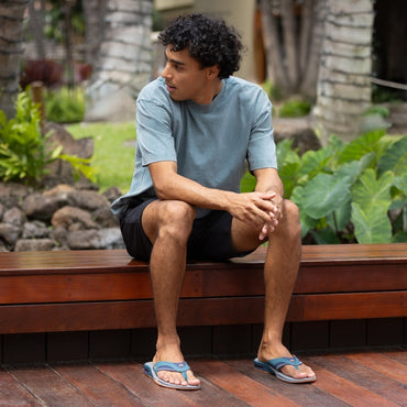 Man wearing Fanning sandals facing forward, seated outdoors on bench, showcasing comfort-focused beach-inspired footwear.
