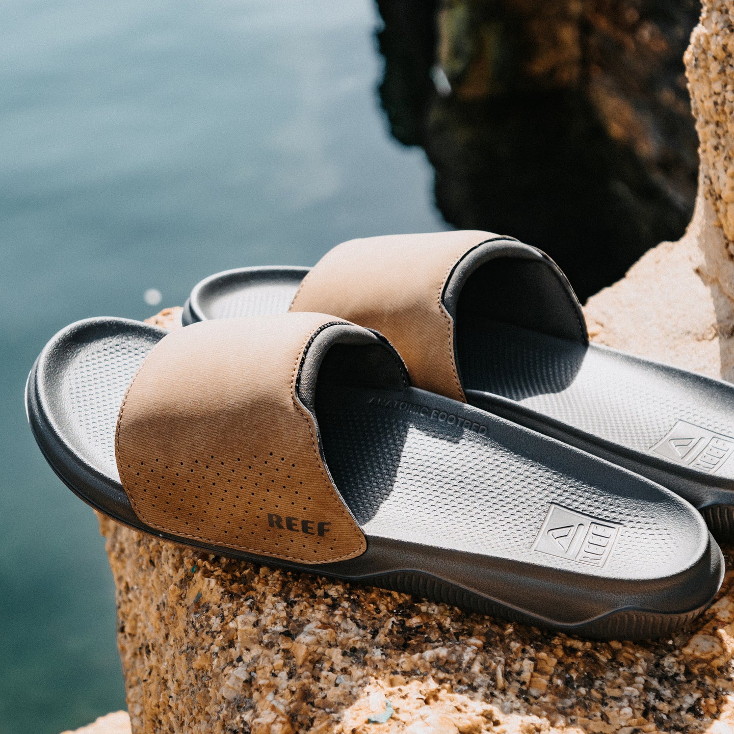 Tailslide tan men's slides facing right with a close up view of padded straps and contoured footbeds on a rocky beach