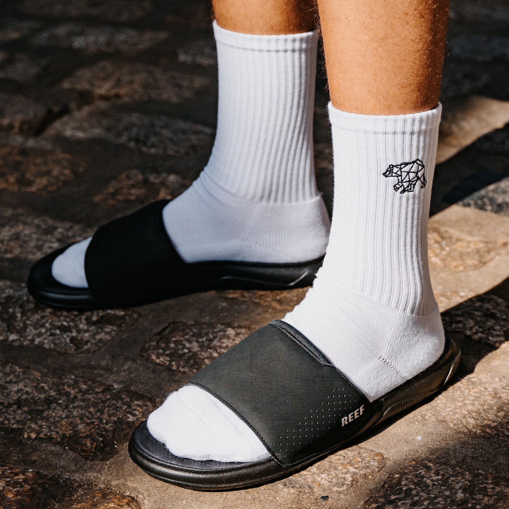 Close-up of black slide sandals facing left, worn with white ribbed socks on a stone surface