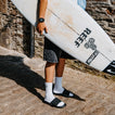 Person carrying a surfboard and wearing black open toe slides with toes facing right on a stone walkway